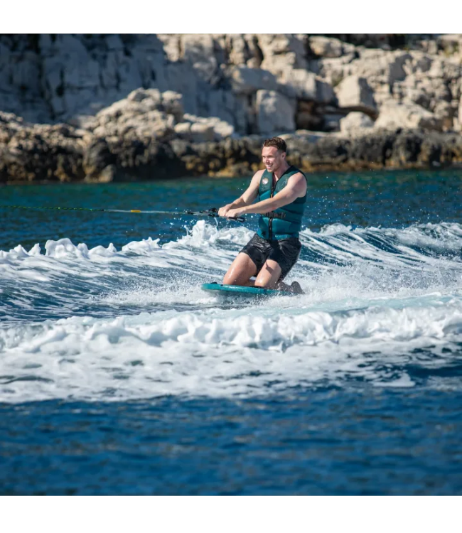 A man wakeboarding on blue water with a rocky coastline in the background, wearing a teal life vest and black shorts.